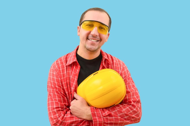 Retrato de un joven ingeniero con un casco en la mano mirando a la cámara sobre un fondo azul.