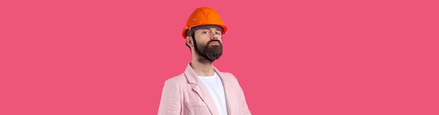 Retrato de un joven ingeniero de construcción que usa un casco naranja en una chaqueta rosa de pie en el fondo rojo del estudio