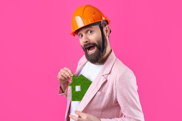 Retrato de un joven ingeniero de construcción que usa un casco naranja en una chaqueta rosa de pie sobre un fondo rojo de estudio Un hombre sostiene una casa ecológica verde