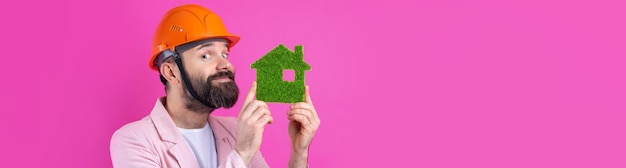 Retrato de un joven ingeniero de construcción que usa un casco naranja en una chaqueta rosa de pie sobre un fondo rojo de estudio Un hombre sostiene una casa ecológica verde