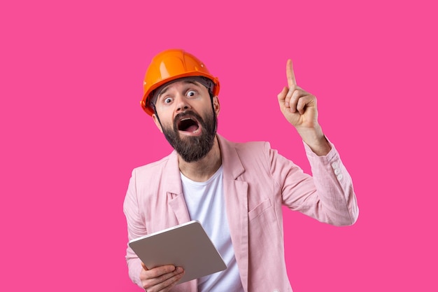 Retrato de un joven ingeniero de construcción que usa un sombrero duro naranja en una chaqueta rosa de pie sobre un fondo rojo de estudio Un hombre con una tableta en las manos