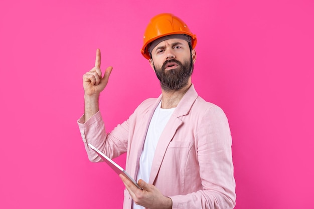 Retrato de un joven ingeniero de construcción que usa un sombrero duro naranja en una chaqueta rosa de pie sobre un fondo rojo de estudio Un hombre con una tableta en las manos