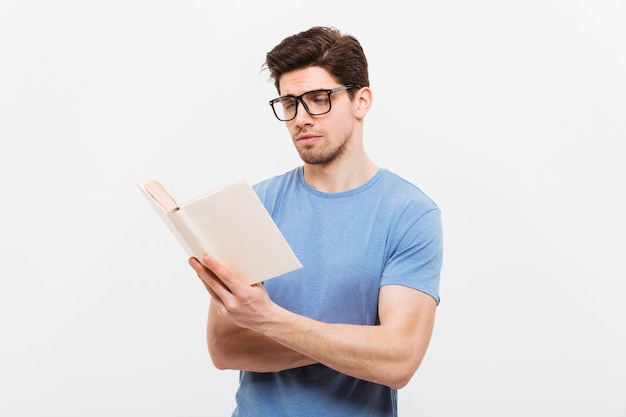 Retrato de joven inteligente en camisa azul con anteojos leyendo libro con concentración, aislado sobre la pared blanca