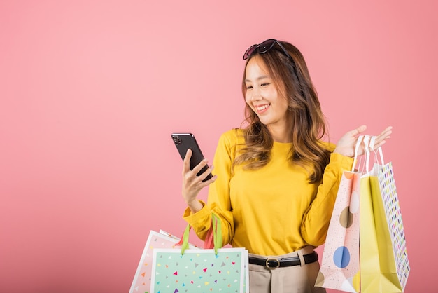 Retrato de una joven joven asiática feliz y hermosa, una adolescente compradora sonriente, de pie emocionada, sosteniendo bolsas de compras en línea, colorida en un teléfono móvil o teléfono inteligente, tomada de estudio aislada.