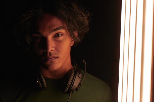 Foto retrato de joven jugador en auriculares mirando a la cámara de pie en una habitación oscura cerca del equipo de iluminación