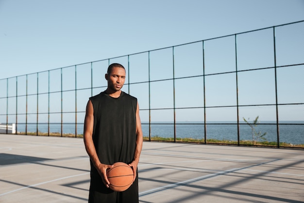 Retrato de un joven jugador de baloncesto de pie en el playgroung y sosteniendo la bola