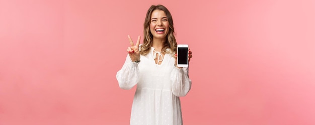 Retrato de una joven kawaii optimista y feliz vestida de blanco que muestra la pantalla del teléfono móvil y el signo de la paz riéndose sintiéndose alegre y contenta de compartir una aplicación increíble enlace útil fondo rosa