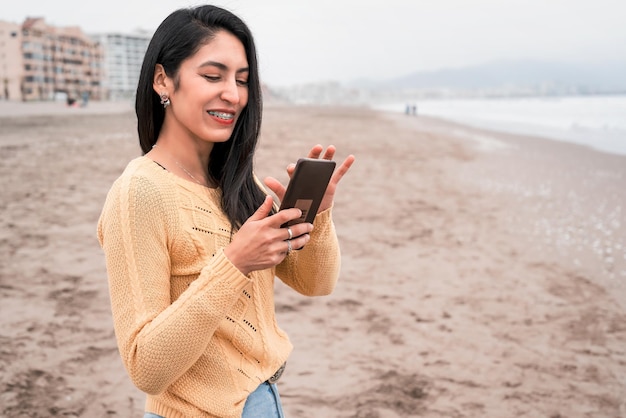 Retrato de una joven latina en la playa sonriendo feliz mirando y desplazándose por el teléfono celular