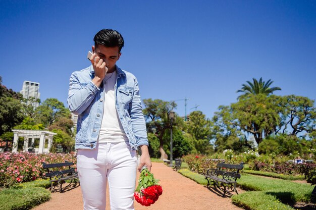 Retrato de un joven latino hispano caucásico con flores en la mano decepcionado llorando después de ver su teléfono caminando en un parque urbano con árboles y cielo en el fondo y copiando espacio