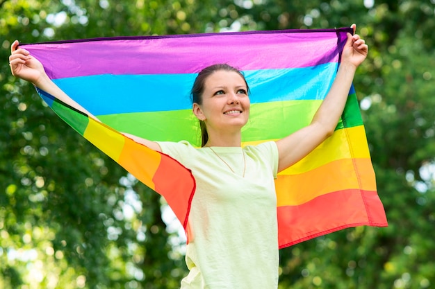 Retrato de joven lesbiana feliz orgullosa mujer, hermosa niña agita la bandera gay de color LGBT del arco iris en un día soleado de verano sobre fondo natural. Comunidad LGBT, concepto de igualdad de relaciones homosexuales