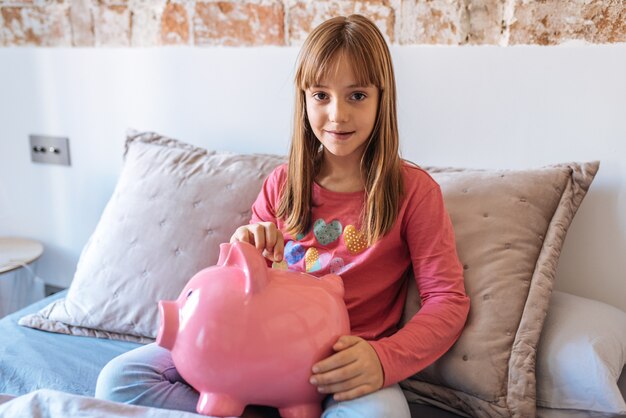 Retrato de una joven linda niña rubia sentada en la cama poniendo dinero en la alcancía