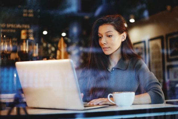 Retrato de joven linda tomando café y usando la computadora portátil en un café
