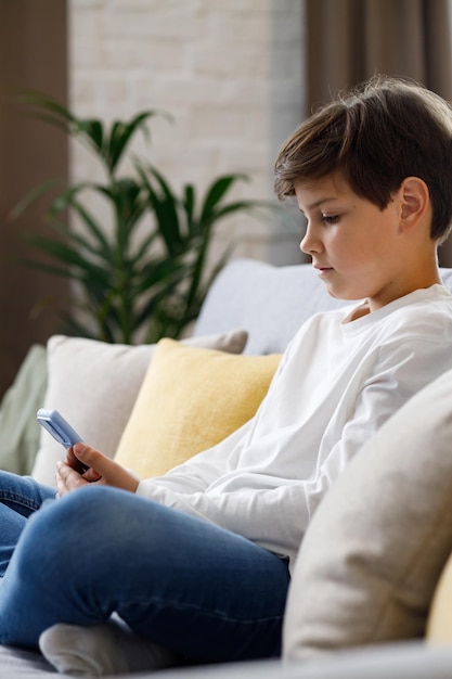 Retrato de un joven lindo sosteniendo un teléfono inteligente sentado en un sofá Adolescente en casa eligiendo música favorita enviando mensajes de texto navegando por Internet viendo videos en las redes sociales jugando juegos en línea