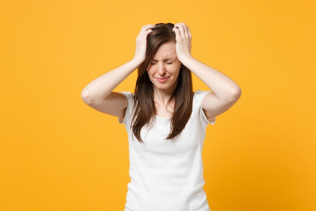 Retrato de una joven llorando disgustada con ropa blanca informal manteniendo los ojos cerrados, poniendo las manos en la cabeza aislada en un fondo naranja amarillo brillante. Concepto de estilo de vida de las personas. Simulacros de espacio de copia.