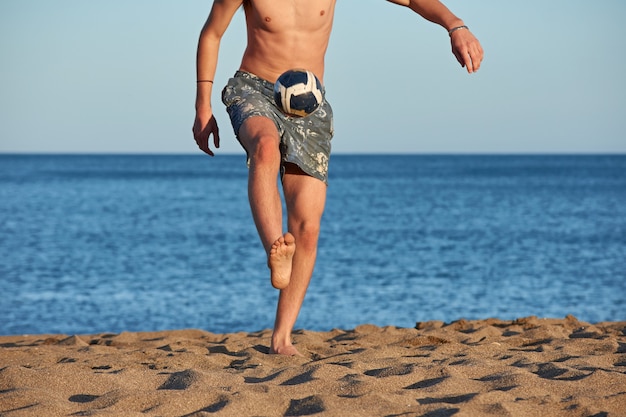 Un retrato de un joven macho caucásico en forma pateando una pelota en la playa