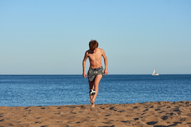 Un retrato de un joven macho caucásico en forma pateando una pelota en la playa