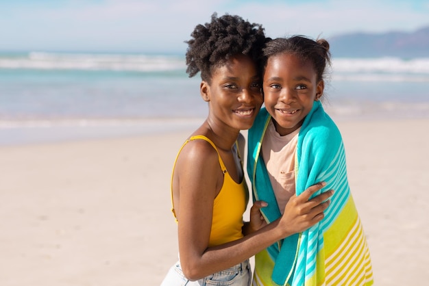 Retrato de una joven madre afroamericana sonriente envolviendo a su hija con una toalla contra el mar y el cielo