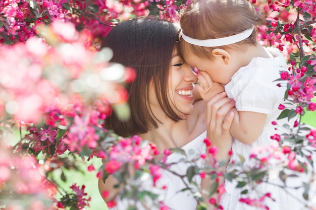 Retrato de joven madre hermosa con su pequeña niña. Todavía cerca de la familia amorosa. Mujer atractiva que detiene a su niño en flores rosadas y la sonrisa.