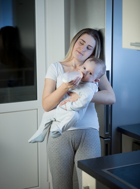 Retrato de joven madre soñolienta alimentando a su hijo del biberón en la cocina por la noche