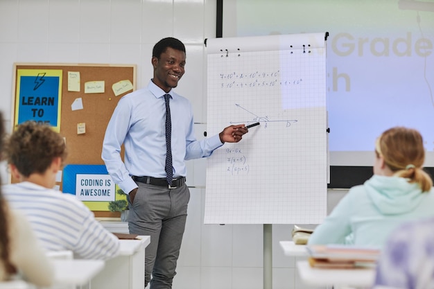 Retrato de un joven maestro negro explicando problemas matemáticos a los niños en clase y sonriendo felizmente poi