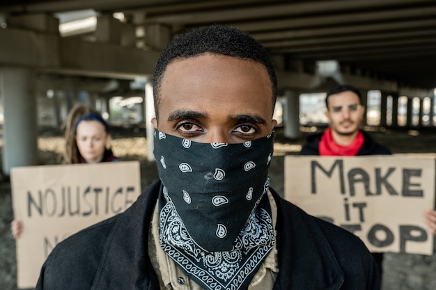 Retrato de joven manifestante negro en pañuelo en la cara de pie contra la gente con carteles debajo del puente