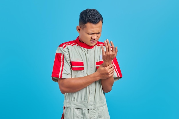 Retrato de joven mecánico asiático que sufre de dolor en las manos y los dedos sobre fondo azul.