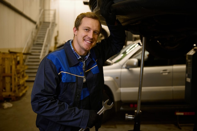 Retrato de un joven mecánico con uniforme profesional, sosteniendo una llave inglesa y trabajando en el servicio de automóviles con un vehículo levantado, sonríe con dientes mirando a la cámara. Reparación y mantenimiento de automóviles.