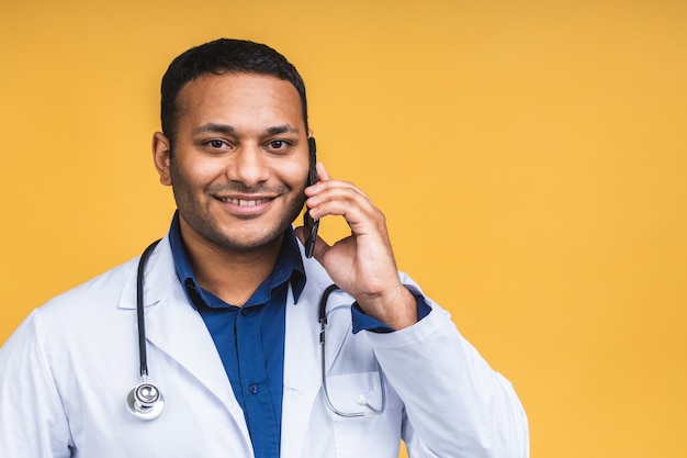 Retrato de joven médico afroamericano hombre negro indio aislado sobre fondo amarillo hablando por teléfono inteligente, sonriendo positivamente. Usando teléfono móvil.