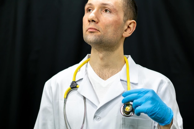 Retrato de un joven médico con una bata blanca sobre un fondo negro Un médico con guantes azules y un estetoscopio médico alrededor del cuello