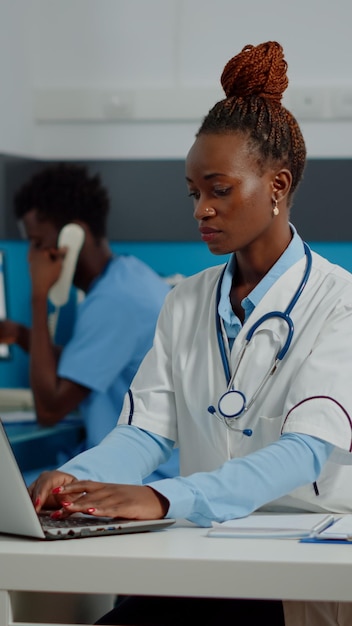 Retrato de un joven médico con bata blanca usando una laptop en un centro de salud. Médico con experiencia profesional escribiendo en un dispositivo de teclado en el escritorio del gabinete médico. Mujer con estetoscopio