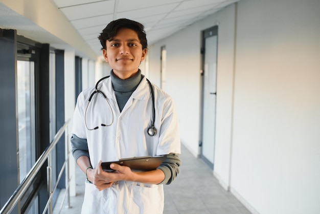 Retrato de un joven médico indio árabe confiado con bata blanca de pie con portapapeles en las manos en el fondo del moderno edificio del hospital al aire libre