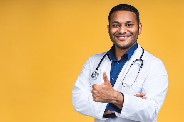 Retrato de joven médico negro indio afroamericano con estetoscopio sobre el cuello en bata médica que se encuentran aisladas sobre fondo amarillo. Pulgares hacia arriba.