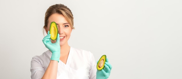 Retrato de joven médico nutricionista con hermosa sonrisa posando a la cámara escondiendo el ojo detrás de medio aguacate sobre fondo blanco, espacio de copia. Beneficios de una nutrición adecuada.