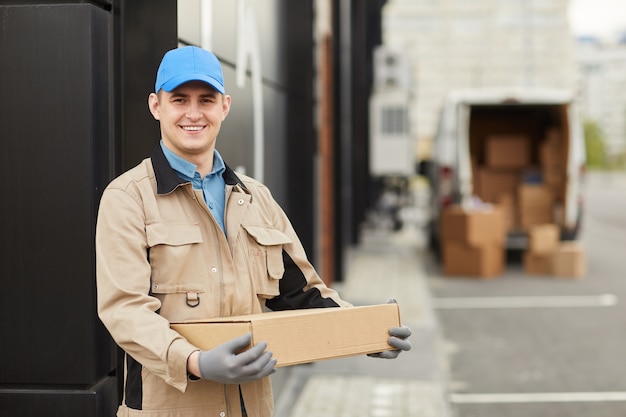 Retrato de joven mensajero en caja de sujeción uniforme y sonriendo a la cámara mientras está de pie al aire libre