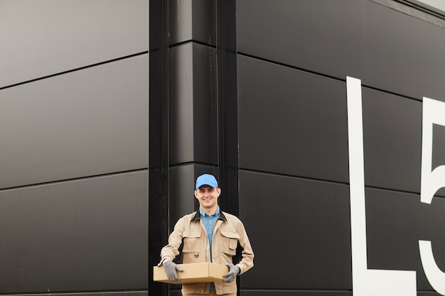 Retrato de joven mensajero en uniforme sosteniendo el paquete y sonriendo a la cámara mientras está de pie contra el almacén al aire libre