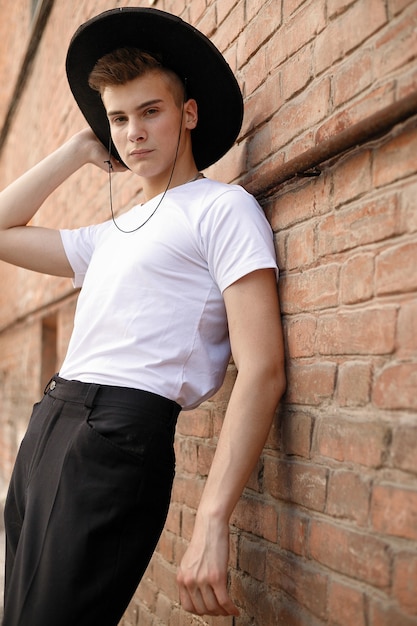 Retrato de un joven de moda en la ciudad. Close-up retrato chico modelo en un sombrero negro un fondo de pared de ladrillo