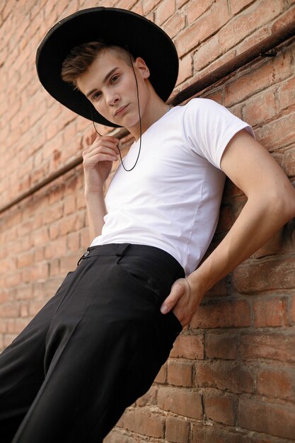 Retrato de un joven de moda en la ciudad. Close-up retrato chico modelo en un sombrero negro un fondo de pared de ladrillo