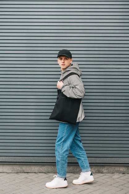 Retrato de un joven de moda en ropa de calle está parado contra una pared gris con una bolsa ecológica negra en la mano