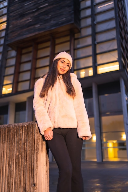 Retrato joven modelo morena sonriente en la ciudad en invierno, vistiendo un suéter y gorro de punto rosa