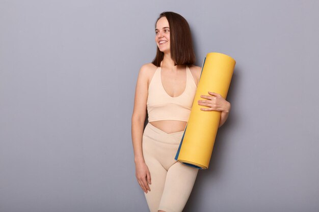 Retrato de una joven modelo que usa ropa deportiva con colchoneta de fitness en las manos mirando hacia otro lado demuestra un cuerpo perfecto haciendo ejercicio en el gimnasio aislado sobre fondo gris