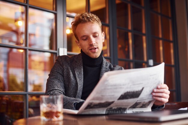 Retrato de joven moderno con ropa formal que se sienta en la cafetería y lee el periódico.