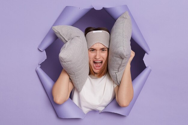 Retrato de una joven molesta, triste y enojada, rompiendo un agujero de papel de fondo morado escuchando un ruido fuerte temprano en la mañana estando de mal humor cubriendo las orejas con almohadas