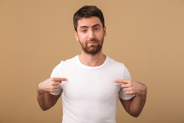 Retrato de un joven molesto vestido casualmente que se encuentran aisladas sobre beige, señalando con el dedo su camiseta en blanco