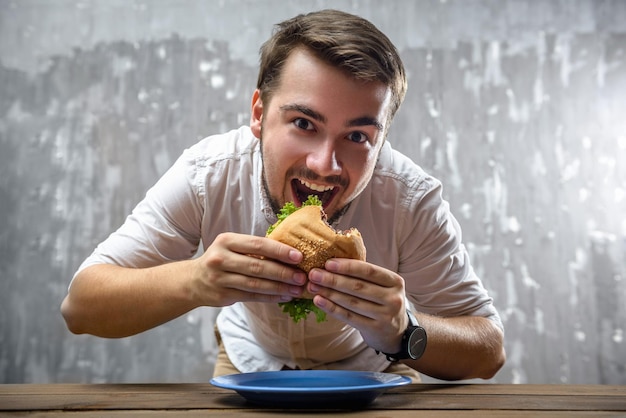 Retrato de un joven mordiendo una hamburguesa con la boca abierta