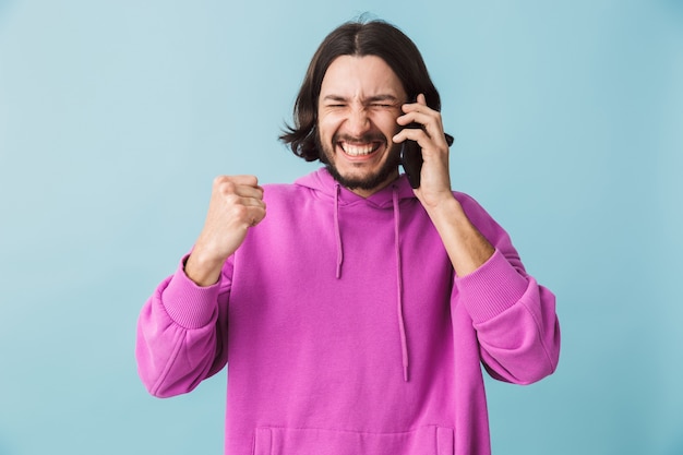 Retrato de una joven morena barbudo hombre vestido con sudadera con capucha que se encuentran aisladas sobre la pared azul, hablando por teléfono móvil, celebrando