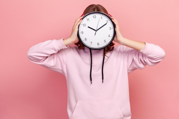 Retrato de una joven morena con capucha sosteniendo un reloj de pared escondiendo su rostro, gestión del tiempo, horario y cita de reunión. Disparo de estudio interior aislado sobre fondo rosa