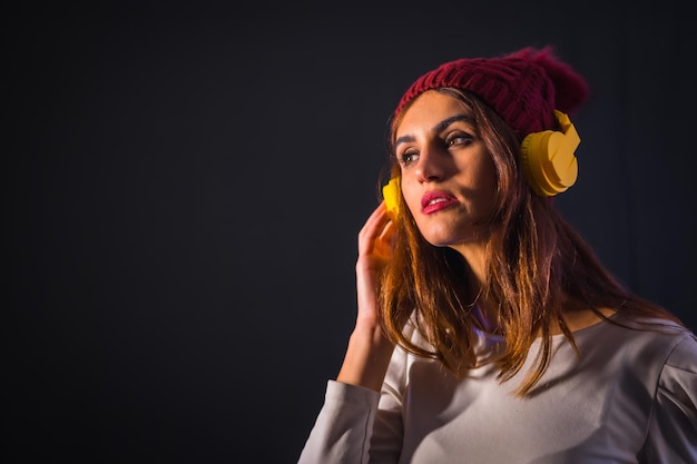 Retrato de una joven morena caucásica con auriculares amarillos, una camiseta blanca y un gorro de lana sobre un fondo negro, concentrado escuchando música