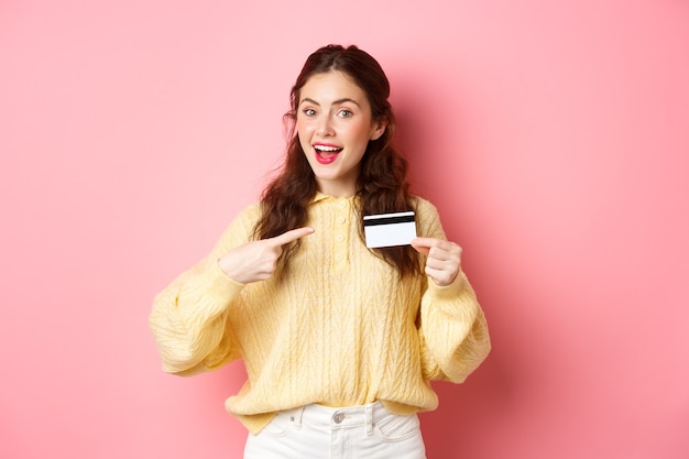 Retrato de joven morena chica caucásica señalando con el dedo a la tarjeta de crédito de plástico recomendando banco ir de compras de pie sobre la pared rosa