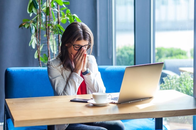 Retrato de una joven morena elegante y deprimida triste con gafas sentada sosteniendo la cabeza hacia abajo y llorando en un estudio interior con fondo de oficina de café