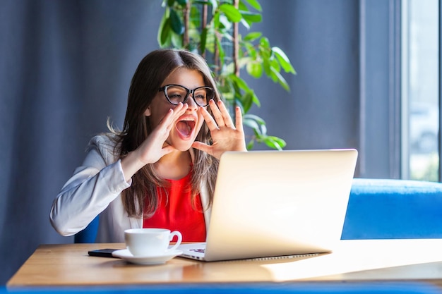 Retrato de una joven morena enojada con anteojos sentada mirando la pantalla de su computadora portátil en una videollamada y gritando para compartir algo importante en el fondo de la oficina del café en el estudio interior